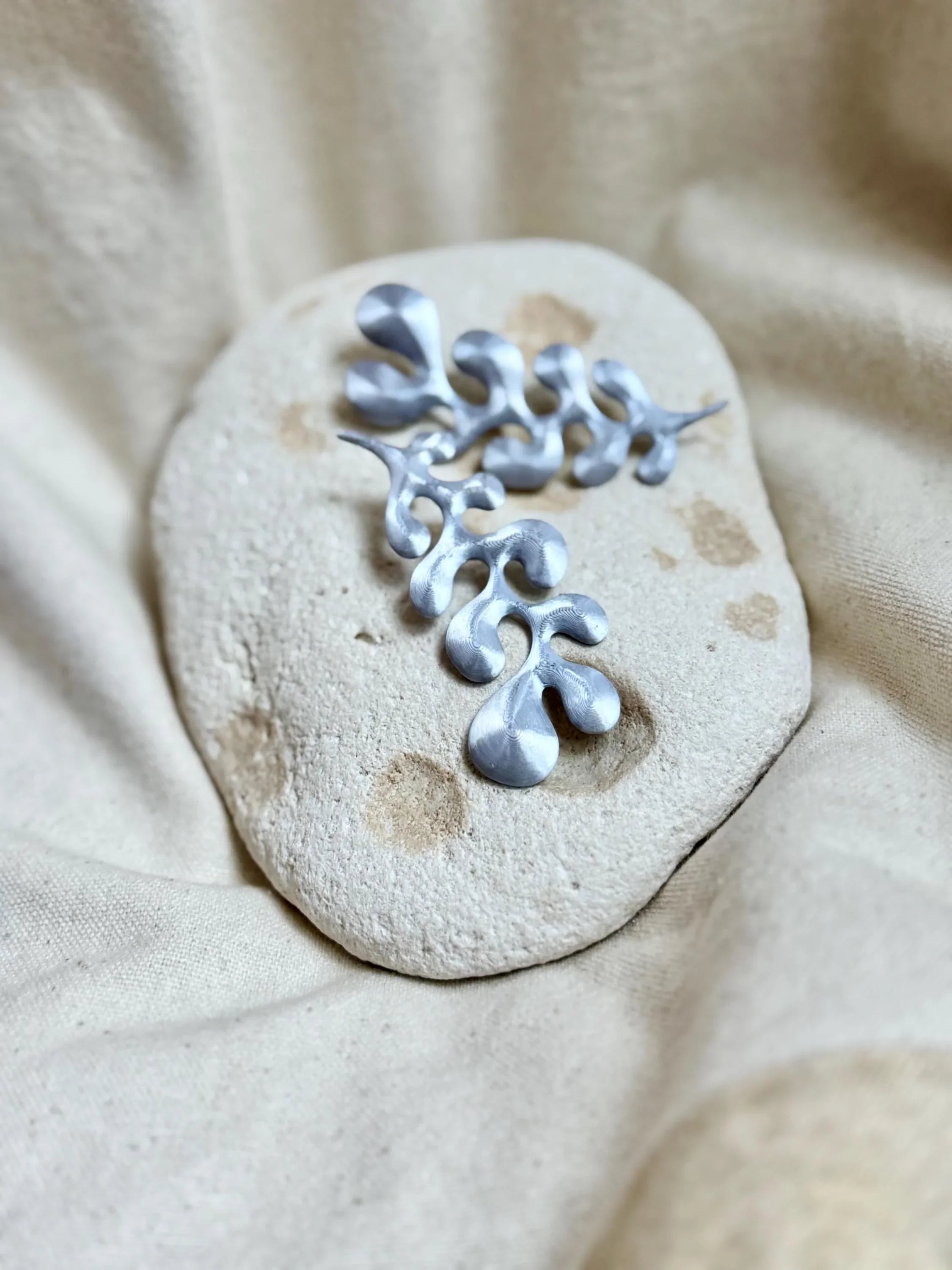 Silver abstract drop-shaped earrings on textured stone with neutral fabric background