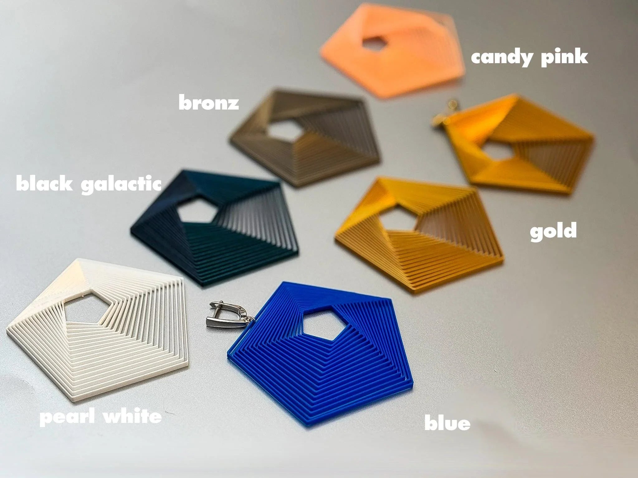 Geometric pentagon earrings in pearl white, blue, black galactic, bronze, candy pink, and gold colors on a flat surface.