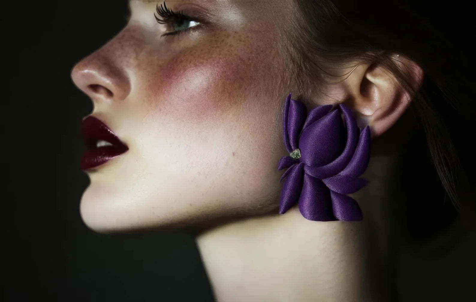 Woman wearing large purple floral statement earring, close-up on ear and face